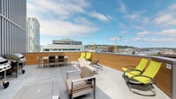 This rooftop terrace is a well-appointed outdoor amenity space designed for both relaxation and social gatherings. The open-air deck features large-format porcelain or concrete tiles underfoot, providing a clean, modern aesthetic that complements the surrounding urban landscape. Warm wood-paneled privacy walls frame the perimeter, adding a natural warmth to the otherwise contemporary design while offering a degree of shelter from the elements.

The space is thoughtfully furnished with a mix of lounging and dining options. Cushioned lounge chairs and sleek chaise lounges are arranged to maximize sun exposure and views, while a dedicated dining area with a table and chairs invites al fresco meals. Pops of yellow-green accent the seating, lending a vibrant, energetic character to the terrace.

From the rooftop, expansive views of the surrounding cityscape unfold, showcasing a dynamic urban skyline with mid-rise buildings, hillside neighborhoods, and active construction cranes that hint at the area's continued growth. The open sky above floods the space with natural light, making it an inviting retreat on clear days. Overall, this rooftop terrace strikes a balance between refined design and relaxed outdoor living, offering an elevated escape above the city.