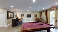 Located at 3801 Glen Verde Trl in Raleigh, NC, this single-floor community clubhouse is designed to provide a comfortable and inviting gathering space for residents. The space is finished with warm, neutral-toned walls, crown molding, and large-format tile flooring throughout, creating a cohesive and polished aesthetic.

The entry lobby sets a welcoming tone, featuring recessed lighting, framed artwork, a dark wood console table with decorative accents, and comfortable seating. A bulletin board and fire alarm control panel are also present, reflecting the facility's functional purpose.

The centerpiece of the clubhouse is a spacious billiards room, anchored by a full-size pool table with a rich burgundy felt surface, set atop a striped area rug. A wall-mounted flat-screen television is positioned for easy viewing, and framed artwork decorates the walls. Double glass doors on one side flood the room with natural light and provide a view of the surrounding greenery outdoors.

An adjacent lounge nook offers a more intimate setting, furnished with colorful upholstered bar-height chairs and a round bistro table beneath a window dressed with striped and red drapery panels. A wall-mounted television and additional framed artwork complete the space. Comfortable armchairs are distributed throughout, encouraging relaxed conversation and leisure throughout the clubhouse.