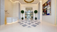 Located in Coral Gables, FL, this grand lobby at Regency Parc is a masterfully designed entrance space that commands attention through its impressive scale and refined aesthetic. The centerpiece of the lobby is its striking geometric patterned marble floor, composed of contrasting white and grey tiles arranged in an intricate diamond and chevron motif that extends throughout the entire expanse. Soaring ceilings amplify the sense of grandeur, while floor-to-ceiling steel-framed windows and monumental glass entrance doors flood the space with natural light and frame views of the lush surroundings outside. The lobby is anchored by a sleek white concierge reception desk, elegantly backlit and set against a textured accent wall bearing the Regency Parc branding. Curated large-format artworks are strategically positioned throughout, adding vibrant color and a gallery-like character to the pristine white walls. Seating areas furnished with deep navy blue curved sofas and accent chairs provide inviting gathering spaces. Manicured topiary trees introduce organic warmth, while a long, light-filled corridor lined with architectural pilasters leads deeper into the building. Recessed and cove lighting throughout ensures a warm, sophisticated ambiance at every hour.