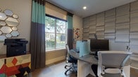 This single-floor business center offers a well-appointed and functional workspace designed to meet a variety of professional needs. The main work area features multiple ergonomic workstations equipped with desktop computers, separated by sleek frosted glass partitions to provide privacy and focus. A large window dressed with two-tone teal and gray curtains allows natural light to fill the room while offering views of the surrounding greenery, creating a bright and comfortable working environment. An accent wall with textured, padded gray panels adds a modern, polished aesthetic to the space.

Adjacent to the workstations, a dedicated printing and utility nook houses a multifunction printer atop a vibrant, geometric-patterned cabinet, complemented by a decorative hexagonal mirror arrangement on the wall. A glass-paneled door provides a clear sightline into a neighboring lounge or game room, hinting at additional amenity spaces nearby.

The business center also benefits from direct access to an expansive outdoor pool area, visible through sliding glass doors. The pool deck is lined with lounge chairs and umbrella-shaded seating, offering a relaxing retreat just steps away. The surrounding stucco exterior with dark wood accents and covered entry points frames the transition between indoor productivity and outdoor relaxation seamlessly.