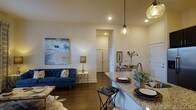 This single-floor residence showcases a well-appointed open-concept layout that effortlessly connects the kitchen and living area, creating a cohesive and inviting atmosphere. The kitchen features a striking granite countertop island with pendant lighting overhead — including a decorative textured glass fixture — providing both a functional prep surface and a casual dining space with bar seating. Rich dark hardwood flooring runs throughout, grounding the space with warmth and contrast against the soft neutral wall tones. Recessed lighting complements the pendant fixtures, ensuring the space is bright and welcoming throughout. The living area is anchored by a plush, tufted navy blue sofa, a cowhide area rug, and a bold abstract wall painting that adds a sophisticated, artistic focal point. A stylish side table with a contemporary lamp adds an elegant finishing touch. The open hallway visible from the main living area suggests additional rooms beyond, contributing to a sense of depth and functionality. Overall, the space balances modern design elements with a comfortable, livable aesthetic, making it both visually appealing and highly practical for everyday living.