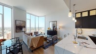 This refined high-rise residence offers a seamless blend of modern design and luxurious finishes throughout its single-floor layout. The open-concept living area is anchored by dramatic floor-to-ceiling corner windows that flood the space with natural light and frame breathtaking panoramic city views, creating a striking backdrop for everyday living. Light hardwood flooring flows throughout, complementing the neutral, contemporary palette.

The gourmet kitchen is a standout feature, showcasing rich dark cabinetry that extends to the ceiling with glass-front upper panels, stainless steel appliances including a gas range with a stainless hood, and an expansive marble-veined quartz island and backsplash. Pendant lighting adds a warm, sophisticated touch. The kitchen opens effortlessly to the living and dining areas, making it ideal for entertaining.

The primary bedroom enjoys its own floor-to-ceiling window with commanding city views and is complemented by an en-suite bathroom featuring large-format marble tile flooring, a glass-enclosed walk-in shower, and refined fixtures. A second bedroom provides a serene retreat with ample closet space. The second bathroom mirrors the same upscale aesthetic, with a glass shower enclosure, dark vanity cabinetry, and crisp marble-style tile throughout. The overall ambiance is polished, airy, and urbane.