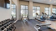 Located at 815 Avenue Plymouth in Mont-Royal, Quebec, this well-appointed fitness facility offers a generously sized, single-floor exercise room designed for a comprehensive workout experience. The space is defined by its contemporary architecture, featuring elevated ceilings with recessed and track lighting that create a bright, energizing atmosphere. Large, floor-to-ceiling windows line the exterior walls, flooding the interior with natural light and providing open views of the surrounding building complex.

The exercise room is finished with warm wood-look flooring across the main cardio and open areas, transitioning to rubberized flooring in the dedicated strength training zones for added safety and functionality. Cardio equipment includes treadmills, elliptical trainers, stationary bikes, and a rowing machine, while the strength area features a dumbbell rack, adjustable benches, a barbell station, and cable machines. Stability balls and resistance accessories are neatly organized on wall-mounted racks.

A distinctive textured white partition wall anchors the central layout, adding a refined architectural detail to the space. Full-length mirrors expand the visual depth of the room, while a wall-mounted flat-screen TV provides entertainment. A glass-partitioned area adjacent to the main floor appears to connect to a separate lounge or amenity space, enhancing the overall facility offering.