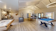 Located at 3555 N First Ave in Tucson, Arizona, this single-floor community clubhouse is a well-appointed recreational and social space designed for both leisure and entertainment. The open-plan layout is anchored by dramatic vaulted ceilings with exposed white beams, bathed in natural light from large glass doors that offer views of the surrounding outdoor area. Two elegant circular chandelier fixtures and track lighting illuminate the space with a warm, inviting glow.

The main game room area is generously sized, housing a regulation pool table, ping pong table, foosball table, and a shuffleboard table, providing a diverse range of recreational options. A cozy lounge corner features leather seating arranged around a tiled fireplace with a wall-mounted television, adding a relaxed gathering spot within the room. A rustic sliding barn door adds a charming architectural detail connecting areas of the space.

The kitchen features a striking geometric-patterned tile backsplash, sleek gray stone countertops, a bar-height island with gold bar stools, light wood cabinetry, and a wall-mounted TV — ideal for social gatherings. Light wood-look flooring flows throughout, unifying the entire space. Curated wall art and lush potted plants complete the modern, resort-inspired aesthetic.