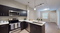 Located at 5428 W Black Hills Ln in Herriman, Utah, this single-floor residence offers a well-designed layout with modern finishes and a clean, contemporary aesthetic. The kitchen is a standout feature, equipped with dark espresso-toned cabinetry, white quartz countertops, and a full suite of stainless steel appliances including a French door refrigerator, electric range with over-the-range microwave, and dishwasher. A peninsula with an undermount sink and pendant lighting above creates an open, functional workspace that flows seamlessly into the living area. Track lighting overhead adds an additional layer of illumination to the kitchen space. Light wood-look flooring runs throughout the main living areas, providing a warm contrast to the darker cabinetry. The living area is open and airy, enhanced by neutral warm-toned walls and natural light from window treatments with horizontal blinds. The bathroom features a white shaker-style vanity with a rich brown countertop, an undermount sink, modern brushed nickel fixtures, and a backlit mirror that provides bright, even lighting. The additional room offers light hardwood-style flooring, neutral beige walls, and a window that allows natural light to filter in, making it a versatile space adaptable to various uses.