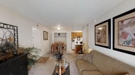 This single-floor residence offers a thoughtfully designed open-concept layout that seamlessly connects the living, dining, and kitchen areas. The living room is a welcoming space, featuring neutral-toned carpeting, warm wall colors, and ample room for comfortable furnishings. Framed wall art and decorative accents contribute to the cozy, inviting ambiance throughout.

The dining area sits adjacent to the living room, providing a defined yet open space for meals and gatherings, with room for a full dining set. A coat closet conveniently positioned near the entryway adds practical storage.

The kitchen is visible from the main living area and features warm wood-toned cabinetry, a breakfast bar or peninsula that bridges the kitchen and dining space, and white appliances. The layout is efficient and well-organized, making the most of the available space.

Natural light filters through the space, complemented by ceiling-mounted light fixtures that keep the interior bright and airy. The consistent neutral color palette of soft whites and beiges throughout creates a clean, cohesive aesthetic. The open flow between rooms enhances the sense of spaciousness, making this residence a functional and comfortable place to call home.