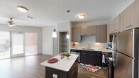 This one-bedroom, one-bathroom apartment in Conroe, TX offers a thoughtfully designed single-story layout with a contemporary aesthetic throughout. The open-concept kitchen serves as the heart of the home, featuring two-tone cabinetry with dark lower cabinets and lighter upper cabinets, quartz countertops, a central island with pendant lighting, and a full suite of stainless steel appliances including a refrigerator, range, microwave, and dishwasher. Under-cabinet lighting adds both functionality and ambiance to the cooking space. A built-in shelving unit adjacent to the kitchen provides convenient storage and display space. The living area is bathed in natural light from large windows with blinds, and durable wood-style plank flooring runs consistently throughout the main living spaces, adding warmth and cohesion. The bedroom is carpeted and accessible via a clean, open doorway from the living area. The bathroom is well-appointed with a full tub and shower combination featuring white subway tile surround, a wood-toned wall cabinet, and modern fixtures. Additional storage is available through a built-in closet with multiple shelves. The unit's neutral color palette of whites and grays creates a clean, move-in-ready atmosphere.