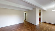 This single-floor residential unit offers a well-organized layout with a variety of functional spaces throughout. The main living area makes a strong first impression with rich hardwood flooring, distinctive exposed ceiling beams set against a textured white ceiling, and neutral cream-toned walls that create a bright, open atmosphere. A large window with vertical blinds allows natural light to fill the space, accompanied by a wall-mounted air conditioning unit and baseboard heating throughout.

The kitchen is efficiently laid out in a galley style, featuring dark wood cabinetry both above and below stone-patterned countertops, a stainless steel sink, a refrigerator, and a dishwasher — providing ample storage and workspace.

The bedrooms are carpeted for comfort, each with windows that bring in natural light and baseboard heating units along the walls. One bedroom includes direct access to a walk-in closet as well as an adjoining bathroom, offering a practical private suite configuration.

The bathroom is clean and functional, with a rich wood vanity topped with a stone-patterned countertop, a large mirror, and a full shower-tub combination. Louvered cabinet doors and chrome fixtures complete the space. The thoughtful layout and consistent finishes throughout give this unit a cohesive and comfortable character.