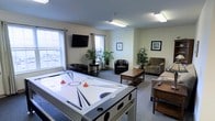 This single-floor recreational lounge is designed to offer a welcoming and functional space for leisure and social interaction. The room's most prominent feature is a full-sized air hockey table positioned at the center, serving as the focal point for entertainment and group activity. Surrounding the game area is a comfortable seating arrangement, including cushioned armchairs and a sofa, all oriented around a wooden coffee table — ideal for relaxed conversation or casual gatherings. A wall-mounted flat-screen television is installed above one of the windows, providing an additional entertainment option for visitors. The space benefits from generous natural light, with multiple windows along the walls fitted with curtains, creating a bright and open atmosphere. The neutral-toned walls and carpeted flooring contribute to a clean, understated aesthetic throughout. Decorative touches such as framed artwork and potted indoor plants are thoughtfully placed around the room, adding warmth and a lived-in character to the environment. The overall layout is open and well-organized, ensuring easy movement throughout the space while accommodating multiple uses simultaneously — from active gameplay to quiet relaxation.