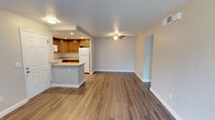 This single-floor unit presents an open-concept design that seamlessly connects the kitchen and living area, creating a spacious and versatile layout. The flooring throughout is a warm-toned wood-look laminate, providing a clean and cohesive aesthetic across both spaces.

The kitchen is equipped with medium-toned wooden cabinetry offering ample storage, granite-look countertops, and a white refrigerator. A peninsula with a raised countertop extends into the living area, serving as both a functional workspace and a casual dining or gathering spot. Recessed lighting in the kitchen provides focused illumination, while white appliances complement the overall neutral palette.

The living area is generously proportioned, featuring light gray walls and a ceiling fan with an integrated light fixture, which enhances airflow and ambient lighting. White baseboards frame the space cleanly throughout. A doorway visible along one wall provides access to additional areas of the unit. The combination of neutral tones and durable flooring gives the space a well-maintained, move-in-ready feel suited for everyday living.