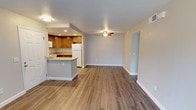 This single-floor unit presents an open-concept design that seamlessly connects the kitchen and living area, creating a spacious and versatile layout. The flooring throughout is a warm-toned wood-look laminate, providing a clean and cohesive aesthetic across both spaces.

The kitchen is equipped with medium-toned wooden cabinetry offering ample storage, granite-look countertops, and a white refrigerator. A peninsula with a raised countertop extends into the living area, serving as both a functional workspace and a casual dining or gathering spot. Recessed lighting in the kitchen provides focused illumination, while white appliances complement the overall neutral palette.

The living area is generously proportioned, featuring light gray walls and a ceiling fan with an integrated light fixture, which enhances airflow and ambient lighting. White baseboards frame the space cleanly throughout. A doorway visible along one wall provides access to additional areas of the unit. The combination of neutral tones and durable flooring gives the space a well-maintained, move-in-ready feel suited for everyday living.
