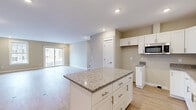 Located at Camden Street in Scarborough, Maine, this 2-bedroom, 1-bathroom single-floor unit offers a thoughtfully designed layout with modern finishes throughout. The open-concept kitchen and living area forms the heart of the home, anchored by a central granite-topped island with shaker-style white cabinetry, brushed nickel hardware, and stainless steel appliances including a built-in microwave. Ample counter space and generous upper and lower cabinet storage make the kitchen both functional and visually appealing. Light-toned hardwood flooring flows seamlessly from the kitchen into the spacious living room, which is bathed in natural light from a large picture window and sliding glass door that opens onto an outdoor balcony with views of the surrounding landscape. Both bedrooms feature the same hardwood flooring, natural light from double-hung windows, and closet storage. The primary bedroom offers a comfortable sense of space. The full bathroom is finished with a granite vanity top, white shaker cabinetry, tile flooring, a tub-shower combination, and a large mirror, all accented by a bright window. Neutral warm-beige wall tones and recessed lighting throughout create a clean, cohesive, and welcoming ambiance.