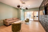 This well-appointed amenity lounge is a thoughtfully designed communal space spread across a single floor, combining modern aesthetics with functional features for residents or occupants. The main lounge area is anchored by a comfortable gray sofa, a sleek white coffee table, and a pair of sage green velvet accent chairs arranged to encourage conversation and relaxation. Light wood-tone flooring runs throughout, complementing the soft teal and warm neutral wall tones that give the space a fresh, inviting atmosphere. A striking multi-arm sputnik-style chandelier with mixed black and brushed gold finishes serves as a focal point overhead, adding a contemporary design element.

Along one wall, a compact dark-cabinetry kitchenette is outfitted with a sink, under-counter refrigerator, and open teal shelving — ideal for casual gatherings. A dramatic textured accent wall clad in layered dark scalloped tiles adds bold visual contrast and artistic depth to the lounge.

A glass-partitioned private room with large windows is accessible from the main lounge, providing a quiet space for focused work or meetings, furnished with a desk and seating. Floor-to-ceiling glass doors open to an outdoor area with views of the surrounding building exterior, bringing natural light deep into the interior. A bank of package lockers is also integrated into the space, adding practical utility.