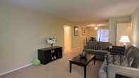 This single-floor residence offers a thoughtfully designed layout that balances comfort and functionality. The living room serves as the heart of the space, featuring plush wall-to-wall carpeting in a neutral tone that complements the warm, light beige walls throughout. A ceiling fan with integrated lighting keeps the area comfortable, while vertical blinds on a large window allow for adjustable natural light, brightening the room throughout the day. The living area is generously sized to accommodate a full seating arrangement, centered around a dark wood coffee table that anchors the space with a grounded, classic aesthetic. A matching dark wood media console adds storage and display space along one wall, adding to the room's cohesive style. Framed wall art contributes a polished, finished look to the interior. The open layout connects the living room directly to the dining area, which features a ceiling light fixture and a dining table set, making it well-suited for casual meals and gatherings. A hallway visible from the living space leads to additional rooms, offering a sense of depth and flow throughout the home. Overall, the space presents a clean, move-in-ready environment with a warm and inviting ambiance throughout.