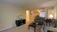 This single-floor residence offers a thoughtfully designed layout that balances comfort and functionality. The living room serves as the heart of the space, featuring plush wall-to-wall carpeting in a neutral tone that complements the warm, light beige walls throughout. A ceiling fan with integrated lighting keeps the area comfortable, while vertical blinds on a large window allow for adjustable natural light, brightening the room throughout the day. The living area is generously sized to accommodate a full seating arrangement, centered around a dark wood coffee table that anchors the space with a grounded, classic aesthetic. A matching dark wood media console adds storage and display space along one wall, adding to the room's cohesive style. Framed wall art contributes a polished, finished look to the interior. The open layout connects the living room directly to the dining area, which features a ceiling light fixture and a dining table set, making it well-suited for casual meals and gatherings. A hallway visible from the living space leads to additional rooms, offering a sense of depth and flow throughout the home. Overall, the space presents a clean, move-in-ready environment with a warm and inviting ambiance throughout.