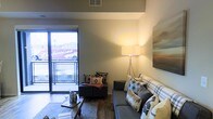 This single-floor urban residence offers a thoughtfully designed living environment that blends contemporary style with comfortable functionality. The living area serves as the heart of the space, featuring warm hardwood-style flooring, neutral-toned walls, and an open layout that promotes an airy, relaxed atmosphere. A large sliding glass door floods the room with abundant natural light and provides direct access to a private balcony, offering open views of the surrounding cityscape — a standout feature that adds both character and connectivity to the urban environment outside. The living area is furnished with a plush sofa accented by a variety of patterned throw pillows, complemented by a sleek coffee table, an additional seating chair, and a modern tripod floor lamp that contributes warm, ambient lighting. A large-format wall art piece adds a bold visual anchor to the space. The ceiling features a recessed HVAC vent, pointing to a modern climate control system integrated seamlessly into the architecture. Overall, the space presents a clean, modern aesthetic with a welcoming ambiance — ideal for those seeking a stylish and functional single-level urban living experience.