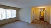This single-floor residential space offers a clean, functional layout designed for comfortable everyday living. The main living area is finished with wall-to-wall carpet in a neutral tone, contributing to a warm and understated ambiance throughout. A large window fitted with vertical blinds allows natural light to filter into the room, brightening the space and offering a view of the surrounding greenery outside. The walls are painted in a soft, neutral palette that enhances the sense of openness and simplicity. An open passageway connects the living area to an adjacent hallway, which leads further into the unit. Warm accent lighting illuminates the connecting corridor, guiding the eye toward additional spaces beyond. Visible from the hallway is what appears to be a compact utility or storage area, adding practical convenience to the layout. The overall design is straightforward and unpretentious, prioritizing livability and ease of use. Clean lines, consistent flooring, and a cohesive color scheme create a unified aesthetic from room to room, making this a welcoming and low-maintenance space well-suited for daily living.