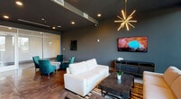 This single-floor space showcases a refined contemporary design, seamlessly blending sophisticated materials with an open, inviting layout. Polished concrete floors run throughout, reflecting ambient and natural light to enhance the spacious feel of the interior. Deep charcoal-toned walls provide a dramatic backdrop, complemented by dark ceilings fitted with recessed lighting that creates a warm, layered glow.

The lounge area is anchored by crisp white sofas and a dark stone coffee table arranged atop a patterned area rug. A bold sputnik-style brass chandelier serves as a striking focal point overhead, while wall-mounted flat-screen televisions add a functional touch. Floor-to-ceiling windows flood this zone with natural light and offer open views of the surrounding outdoor landscape.

A reception or service counter features a dramatic black-and-gold veined marble surface with warm wood cabinetry below, positioned near additional seating clusters of richly upholstered accent chairs. Abstract artwork adorns the walls nearby.

The bar area commands attention with an expansive, deeply veined green marble countertop and herringbone wood-patterned base, surrounded by upholstered bar stools. A textured gold tile accent wall and dual mounted screens complete the backdrop, while pendant lighting overhead adds an elegant finishing detail throughout the space.