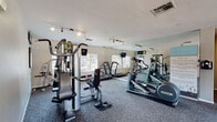 This single-floor fitness center offers a comprehensive workout environment designed to support a wide range of exercise routines. The space is organized into distinct functional zones, providing both cardio and strength training options for users of all fitness levels.

The cardio area is outfitted with multiple treadmills, elliptical machines, and a recumbent stationary bike, all positioned to take advantage of large windows that allow natural light to fill the room and offer views of the surrounding outdoor area. Mounted flat-screen televisions are positioned throughout the space, providing entertainment during workouts.

The strength training section features a multi-station cable machine with instructional charts displayed on the wall, along with an adjustable workout bench and additional resistance equipment. A dedicated TRX Training Zone is clearly marked and equipped with suspension training systems, accompanied by posted exercise guides for easy reference.

Floor-to-ceiling mirrors line one wall, visually expanding the space and allowing users to monitor their form. Track lighting runs along the ceiling, ensuring bright, even illumination throughout. The flooring consists of durable, low-pile carpet suitable for fitness use.

A striking large-scale landscape mural depicting a dramatic canyon sunrise serves as a bold focal point on one wall, adding an energizing visual element to the space. The overall design is clean, functional, and welcoming.
