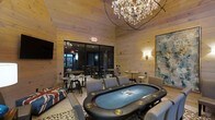 This single-level entertainment lounge is thoughtfully designed for social gatherings and leisure. The centerpiece of the main room is a full-size poker table with built-in cup holders and a blue felt surface, surrounded by plush upholstered seating. Warm, light-toned wood paneling lines both the walls and ceiling, giving the space a cozy yet refined cabin-like character. Elevated ambiance is achieved through a striking crystal chandelier overhead, complemented by elegant wall sconces that cast a soft, inviting glow. A large abstract artwork adorns one wall, adding a contemporary artistic touch to the warm interior. A Union Jack-patterned accent sofa contributes a playful, eclectic personality to the décor. Large sliding glass doors open to an adjoining bar area furnished with high-top tables and industrial-style bar stools, extending the entertainment space and offering a seamless transition between zones. The bar area features additional chandelier lighting, maintaining visual continuity throughout. A wall-mounted flat-screen television provides added entertainment options. Together, the design elements create a sophisticated yet relaxed atmosphere ideal for hosting guests, game nights, and casual socializing.