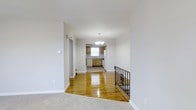 This single-floor space features a thoughtfully arranged open layout that connects the living area directly to the kitchen, creating a cohesive and functional flow. The living area is finished with plush carpeting, while the central and kitchen zones showcase polished hardwood floors that reflect light beautifully, adding warmth and character to the interior. Crisp white walls and white baseboards throughout lend the space a clean, bright aesthetic.

A wrought iron staircase railing with decorative scrollwork adds a subtle architectural detail near the transition between the carpeted and hardwood sections. An elegant multi-bulb chandelier hangs above, serving as a focal point and providing ambient illumination to the central area.

The kitchen is equipped with medium-toned wood cabinetry, a white refrigerator, a dishwasher, and a sink positioned beneath a window that allows natural light to filter in. The kitchen floor features a neutral tile or linoleum surface, distinguishing it from the hardwood in the adjoining space. A doorway off the main living area leads to an additional room, offering practical separation within the layout. Overall, the space presents a tidy, well-maintained environment with classic detailing.