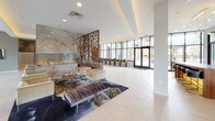 This elegantly designed lobby offers a refined first impression with large-format porcelain tile flooring, soaring ceilings, and an abundance of natural light streaming through expansive floor-to-ceiling windows. At the heart of the space, a dramatic linear fireplace set against a sweeping onyx marble-clad wall creates a striking focal point, framed by tall, sculptural laser-cut wood screens that add warmth and visual depth while subtly dividing zones within the open layout. A generously sized bar-height communal table with sleek leather-and-gold stools sits adjacent to the window wall, offering views of the landscaped exterior streetscape. Recessed tray ceilings with integrated lighting lend a polished architectural detail overhead. A dedicated lounge area features plush velvet seating arranged on a richly patterned area rug, inviting conversation and relaxation. A refreshment station with warm wood cabinetry and dark stone countertops sits near a pair of stainless steel elevators. An enclosed glass-walled conference or leasing office is accessible from the main floor, finished with black steel framing. The entry vestibule features floor-to-ceiling glazing with dark metal framing, sheer blue drapery panels, and a monogrammed entry mat, setting a distinguished tone from the moment of arrival.