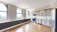 Located at 50 W. Front Street in the heart of downtown Columbus, Ohio, this one-bedroom, one-bathroom residence offers a well-planned layout across a single floor. The open-concept kitchen and living area serves as the centerpiece of the unit, featuring light hardwood flooring, arched windows with sweeping city views, and a bold two-tone accent wall. The kitchen is outfitted with stainless steel appliances including a gas range, microwave, and dishwasher, complemented by gray shaker-style cabinetry and a classic white tile backsplash. A long, well-lit hallway connects the main living area to the private quarters, with clean white walls and recessed lighting that create a bright, welcoming passage. The bedroom is finished with plush wall-to-wall carpeting, neutral warm-toned walls, and a large window framing city views, alongside a spacious closet. The bathroom features polished tile flooring, a dual-sink vanity with gray cabinetry and a white countertop, and a glass-enclosed walk-in shower with a rainfall showerhead. Additional conveniences include an in-unit laundry area. The unit's modern finishes, natural light, and urban vistas make it a refined city residence.