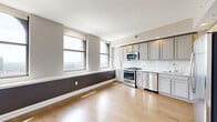 Located at 50 W. Front Street in the heart of downtown Columbus, Ohio, this one-bedroom, one-bathroom residence offers a well-planned layout across a single floor. The open-concept kitchen and living area serves as the centerpiece of the unit, featuring light hardwood flooring, arched windows with sweeping city views, and a bold two-tone accent wall. The kitchen is outfitted with stainless steel appliances including a gas range, microwave, and dishwasher, complemented by gray shaker-style cabinetry and a classic white tile backsplash. A long, well-lit hallway connects the main living area to the private quarters, with clean white walls and recessed lighting that create a bright, welcoming passage. The bedroom is finished with plush wall-to-wall carpeting, neutral warm-toned walls, and a large window framing city views, alongside a spacious closet. The bathroom features polished tile flooring, a dual-sink vanity with gray cabinetry and a white countertop, and a glass-enclosed walk-in shower with a rainfall showerhead. Additional conveniences include an in-unit laundry area. The unit's modern finishes, natural light, and urban vistas make it a refined city residence.
