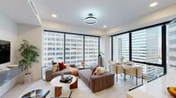 This contemporary high-rise residence is defined by its clean lines, neutral palette, and an abundance of natural light pouring through expansive floor-to-ceiling windows that frame striking urban skyline views throughout. The open-plan living and dining area serves as the heart of the space, anchored by a circular LED ceiling fixture and furnished with a generous sectional sofa, glass-topped coffee table, and a compact dining set — all bathed in city light. The kitchen is thoughtfully designed with two-tone cabinetry in white and warm wood, a white quartz waterfall island with bar seating, stainless steel appliances, and a geometric pendant light fixture overhead. Both bedrooms are bright and spacious, each with floor-to-ceiling windows overlooking the surrounding cityscape. The primary bedroom includes a walk-in closet, while a secondary bedroom offers a versatile layout with a desk area and lounge seating. The primary bathroom features a wide vanity with wood cabinetry, a white quartz countertop, an illuminated mirror, and marble-look tile flooring. A second bathroom mirrors the same refined aesthetic with sleek fixtures and contemporary artwork. Throughout, the residence maintains a cohesive modern sensibility with high-end finishes and an emphasis on light and urban connectivity.