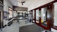 This single-story fitness center is thoughtfully designed to support a comprehensive workout experience. The space is divided into distinct functional zones, beginning with a dedicated cardio area housing multiple treadmills, ellipticals, and a stationary bike, all positioned near windows with wood-trimmed blinds that allow natural light to filter in. Mounted flat-screen televisions are positioned throughout for entertainment during workouts.

The strength training area features a multi-station cable machine, a wall-mounted barbell rack with TRX suspension trainers, free weights including a curl bar, medicine balls, and a weight bench. Full-length mirrors line one wall, visually expanding the space and allowing users to monitor their form. A large wall clock anchors the mirrored section, adding a functional and stylish detail.

The flooring consists of durable gray rubberized material suited for heavy equipment and high-traffic use, with a textured entry mat at the glass-paneled entrance doors framed in rich dark wood. Recessed lighting and a ceiling fan provide comfortable illumination and airflow. The overall aesthetic balances function with a clean, modern design through a neutral color palette accented by dark gray and brown tones.
