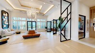 This generously proportioned single-floor space showcases a sophisticated modern design characterized by light hardwood flooring, soaring double-height ceilings, and an abundance of natural light streaming through large black-framed windows and glass doors. The entrance is defined by a dramatic steel-and-frosted-glass partition that creates an elegant visual boundary while maintaining an open, airy feel. A coffered ceiling with warm wood detailing and cascading chandelier fixtures anchors the primary lounge area, where expansive glazing offers outdoor views.

The living and lounge zone features multiple distinct seating arrangements, a built-in media wall with decorative wood-paneled surround, and integrated shelving niches. A statement patterned accent wall highlights a beverage station in one corner, adding a bold visual focal point.

The kitchen is outfitted with warm wood cabinetry, a long quartz-topped island with bar seating, stainless steel appliances, and a sculptural metallic chandelier overhead. Adjacent to the kitchen, an entertainment area with an exposed wood-lattice ceiling houses a billiards table, built-in banquette seating, and a dining zone, creating a seamlessly flowing social environment throughout the space.