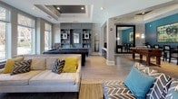 Located at 102 Fallsgrove Blvd in Rockville, Maryland, this single-floor community clubhouse offers a well-appointed and thoughtfully designed gathering space for residents. The open-concept layout connects multiple distinct zones, all unified by warm hardwood-style flooring, tray ceilings with recessed lighting, and a sophisticated neutral color palette accented by teal walls and white trim.

The main lounge area serves as the heart of the space, featuring comfortable seating arrangements centered around a sleek fireplace flanked by large windows that flood the room with natural light and offer views of the outdoor pool area. A wall-mounted flat-screen television above the fireplace makes this an ideal spot for relaxation or group gatherings.

Adjacent to the lounge is a billiards and game room, complete with a full-size pool table and foosball table, providing ample entertainment options. Built-in bookshelves stocked with reading material and framed artwork lend a refined, library-like atmosphere to this area, enhanced further by tray ceiling detailing.

The dining and casual seating areas offer flexible spaces for socializing, complemented by stylish furnishings. The fully equipped kitchen features white shaker cabinetry, dark granite countertops, stainless steel appliances including a side-by-side refrigerator, microwave, and dishwasher, making it ideal for catered events or resident use. Throughout, the space maintains a polished yet inviting ambiance.