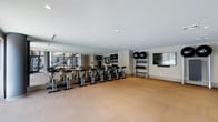 Core Fitness is a purpose-built, single-floor fitness studio designed to support a streamlined and effective workout experience. The space is defined by its clean, modern aesthetic, featuring neutral tones, recessed lighting, and a smooth, cushioned floor surface well-suited for high-intensity training.

A full wall of floor-to-ceiling mirrors lines one side of the studio, amplifying the sense of space and allowing users to monitor their form during exercise. A row of stationary spin bikes is arranged along this mirrored wall, creating a dedicated cycling area ideal for group or individual cardio sessions.

Along the adjacent wall, a multi-tiered equipment rack holds an organized selection of medicine balls and stability balls in varying sizes, providing convenient access to functional training tools. A wall-mounted flat-screen display is positioned nearby, suitable for streaming instructional content or fitness programs.

The recessed ceiling lights provide even, consistent illumination throughout the studio, while the open floor plan allows for flexible use of the central space for stretching, floor exercises, or additional equipment. The overall ambiance is focused and professional, reflecting a space thoughtfully designed for fitness performance.