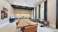 This single-floor workspace is thoughtfully divided into three distinct areas, each designed to support different working styles. The main collaborative zone centers around a substantial solid-wood communal table with leather rolling chairs, set against a wall of large windows dressed with floor-to-ceiling charcoal drapes that frame views of surrounding greenery. Curated artwork lines the walls alongside a mounted flat-screen display, adding a creative, gallery-like atmosphere. A dedicated workstation area features a built-in white desk running along a striking three-dimensional hexagonal wood-tile accent wall, illuminated by industrial-style pendant lights overhead — offering individual seating for focused, heads-down work. A private meeting room provides an intimate setting with a round walnut table, upholstered accent chairs, a wall-mounted television, and a textured chevron-patterned accent wall, ideal for small group discussions or presentations. Throughout, neutral-toned carpet, recessed lighting, and a consistent modern aesthetic tie the spaces together cohesively, creating a professional yet inviting environment suited for productivity and collaboration.
