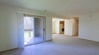 This single-floor residence offers a thoughtfully arranged layout with an open and airy feel throughout. The main living area is bright and spacious, featuring wall-to-wall carpeting and a large sliding glass door that fills the room with natural light and provides direct access to an outdoor deck. The deck overlooks a lush, green yard with mature trees, creating a pleasant indoor-outdoor connection. A ceiling fan is positioned at the transition point between the living area and an adjoining room, enhancing airflow and comfort throughout the connected spaces. The kitchen is visible from the main living area and is equipped with standard appliances, including a refrigerator and range, along with warm-toned cabinetry that adds a classic touch. The open sightlines between the living area, adjoining room, and kitchen give the home a cohesive and functional flow. Neutral wall tones and light-colored carpeting throughout contribute to a clean, understated aesthetic that is easy to personalize. The layout is well-suited for everyday living, with practical connections between the key areas of the home.