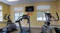 This fitness center offers a practical and comfortable space designed to support an active lifestyle. The room is finished with warm yellow walls and carpeted flooring, contributing to a welcoming atmosphere. Multiple windows line the walls, allowing ample natural light to fill the space and providing views of the surrounding greenery outdoors — an inviting backdrop for any workout session. A wall-mounted flat-screen television is centrally positioned, allowing users to stay entertained while exercising. The facility is equipped with a selection of cardio machines, including a treadmill and an elliptical trainer, along with additional exercise equipment visible throughout the room. The single-floor layout keeps everything accessible and easy to navigate. The overall design balances functionality with comfort, making it a convenient and well-appointed space for daily fitness routines.