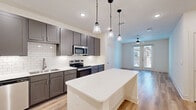 This well-designed one-bedroom, one-bathroom unit in Denton, TX offers a thoughtfully planned open-concept layout with consistent, modern finishes throughout. Light wood-look flooring runs throughout the entire space, contributing to a clean, cohesive aesthetic.

The kitchen is a standout feature, showcasing grey shaker-style cabinetry, a classic white subway tile backsplash, white quartz countertops, and a full suite of stainless steel appliances including a refrigerator, electric range, microwave, and dishwasher. A central island with pendant lighting above provides additional prep space and a natural focal point connecting the kitchen to the living area.

The living area is bright and open, benefiting from natural light provided by French doors that lead to an outdoor balcony. A ceiling fan adds comfort, and the clean white walls create a versatile backdrop.

The bedroom is a comfortable size, featuring two windows that allow ample natural light and views of the outdoors, along with its own ceiling fan. It connects directly to a spacious walk-in closet with built-in shelving and hanging rods, as well as to the full bathroom.

The bathroom is finished with a dual-sink vanity featuring dark cabinetry and white countertops, a subway tile surround tub-and-shower combination, and recessed lighting, presenting a clean and functional design.