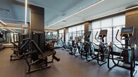 Located at 1000 Schindler Dr in South Amboy, NJ, this modern fitness center is designed to deliver a premium workout experience. The space is thoughtfully laid out across a single floor, maximizing usability while maintaining an open, spacious feel. Large windows line the perimeter, flooding the interior with abundant natural light and offering views of the surrounding building exterior, creating an energizing atmosphere for exercise.

The fitness center features a robust selection of cardio equipment, including a full row of treadmills, elliptical machines, and stationary and spin bikes, all equipped with individual digital displays for performance tracking. Complementing the cardio zone, a dedicated strength training area houses cable machines, a functional training rig, and weight benches to support a full range of resistance workouts.

The interior design is sleek and contemporary, characterized by a dark, speckled rubber flooring that provides durability and comfort underfoot. Linear LED lighting runs along the ceiling, casting a clean, even glow throughout the space. Floor-to-ceiling mirrors on select walls enhance the sense of depth and allow users to monitor their form. Entry is through glass-paneled double doors, and a decorative mural accent wall adds a distinctive visual element. The overall ambiance is polished, functional, and well-suited for serious fitness enthusiasts.