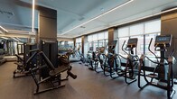 Located at 1000 Schindler Dr in South Amboy, NJ, this modern fitness center is designed to deliver a premium workout experience. The space is thoughtfully laid out across a single floor, maximizing usability while maintaining an open, spacious feel. Large windows line the perimeter, flooding the interior with abundant natural light and offering views of the surrounding building exterior, creating an energizing atmosphere for exercise.

The fitness center features a robust selection of cardio equipment, including a full row of treadmills, elliptical machines, and stationary and spin bikes, all equipped with individual digital displays for performance tracking. Complementing the cardio zone, a dedicated strength training area houses cable machines, a functional training rig, and weight benches to support a full range of resistance workouts.

The interior design is sleek and contemporary, characterized by a dark, speckled rubber flooring that provides durability and comfort underfoot. Linear LED lighting runs along the ceiling, casting a clean, even glow throughout the space. Floor-to-ceiling mirrors on select walls enhance the sense of depth and allow users to monitor their form. Entry is through glass-paneled double doors, and a decorative mural accent wall adds a distinctive visual element. The overall ambiance is polished, functional, and well-suited for serious fitness enthusiasts.