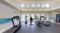 Located at 150 Arbors Cir in Gahanna, Ohio, this well-equipped fitness center offers a bright and functional environment designed to support a comprehensive workout routine. The space is characterized by soaring ceilings in the cardio zone, where a row of large transom windows and full-length glass doors flood the room with natural light and provide open views of the surrounding greenery. A distinctive teal accent stripe runs along the perimeter walls, lending the space a clean, modern aesthetic against the predominantly white interior. The durable rubber flooring throughout provides excellent support and traction for a variety of exercises. The cardio area is outfitted with multiple treadmills, elliptical trainers, and stationary bikes, all positioned to take advantage of the natural light and outdoor views. A separate strength training zone features a cable machine, adjustable bench, and wall-mounted accessories, with floor-to-ceiling mirrors that visually expand the space and allow for proper form checking during workouts. Recessed and flush-mounted lighting ensures the facility is well-lit throughout. A hallway connects to additional amenities, and a beverage vending machine is conveniently accessible near the entrance. The overall ambiance is clean, energizing, and well-maintained, making it an inviting space for daily fitness.