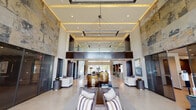Tradehouse presents a striking single-story clubhouse and leasing facility defined by its dramatic architectural design and upscale contemporary interiors. The grand lobby commands immediate attention with its soaring double-height ceilings, coffered tray detailing with warm amber cove lighting, and an expansive stone accent wall that rises the full height of the space. A geometric pendant chandelier anchors the room, while polished concrete floors and floor-to-ceiling glass walls flood the interior with natural light and frame views of the surrounding landscaping and outdoor pool area.

Carefully arranged seating vignettes throughout the lobby invite guests to relax, featuring tailored sofas, accent chairs with layered textiles, and sculptural decorative objects. The Tradehouse reception desk is finished in stone and dark wood, accented by elegant pendant lighting suspended from above. Adjacent to the main desk, a well-appointed service counter features textured wave-pattern tile, stone countertops, stainless steel appliances, and a coffee station.

Additional lounge areas include intimate conversation spaces with sectional seating, large-scale windows overlooking the pool, and refined built-in lighting. Private offices and corridors extend from the main areas, maintaining the cohesive modern aesthetic throughout with dark-framed glass partitions and rich wood tones.
