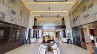 Tradehouse presents a striking single-story clubhouse and leasing facility defined by its dramatic architectural design and upscale contemporary interiors. The grand lobby commands immediate attention with its soaring double-height ceilings, coffered tray detailing with warm amber cove lighting, and an expansive stone accent wall that rises the full height of the space. A geometric pendant chandelier anchors the room, while polished concrete floors and floor-to-ceiling glass walls flood the interior with natural light and frame views of the surrounding landscaping and outdoor pool area.

Carefully arranged seating vignettes throughout the lobby invite guests to relax, featuring tailored sofas, accent chairs with layered textiles, and sculptural decorative objects. The Tradehouse reception desk is finished in stone and dark wood, accented by elegant pendant lighting suspended from above. Adjacent to the main desk, a well-appointed service counter features textured wave-pattern tile, stone countertops, stainless steel appliances, and a coffee station.

Additional lounge areas include intimate conversation spaces with sectional seating, large-scale windows overlooking the pool, and refined built-in lighting. Private offices and corridors extend from the main areas, maintaining the cohesive modern aesthetic throughout with dark-framed glass partitions and rich wood tones.