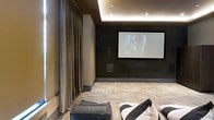 This dedicated home theater room is designed with both function and sophistication in mind, offering a purpose-built environment for an immersive entertainment experience. The focal point of the room is a large wall-mounted projection screen set against a deep, dark-toned feature wall, which enhances contrast and minimizes light reflection during viewing. Flanking the screen, in-wall speakers are integrated seamlessly into the design, supporting a high-quality surround sound setup.

The ceiling features elegant cove lighting that emits a warm, indirect ambient glow, adding a cinematic atmosphere while providing subtle illumination without compromising screen visibility. The ceiling itself is finished with a recessed tray design, contributing to the room's refined aesthetic.

Floor-to-ceiling drapes in a rich, textured fabric line one wall, adding acoustic dampening properties alongside a luxurious visual element. The flooring consists of patterned carpet tiles in neutral tones, which further aid in sound absorption. Comfortable seating, including a plush sofa with decorative pillows, completes the space, ensuring a relaxed and inviting viewing environment. The overall ambiance is dark, intimate, and polished.