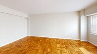 Unit 712 at 35 E St NW in Washington, DC offers a efficiently designed single-floor studio layout in the heart of the city. The main living area is characterized by warm parquet hardwood flooring arranged in a classic basket-weave pattern, neutral beige walls, and white trim throughout. A large window along one wall admits natural light and houses a wall-unit air conditioner, complemented by a radiator below for year-round climate control.

The kitchen is set apart in its own defined alcove, featuring rich dark wood cabinetry — including upper glass-front display cabinets — dark countertops, a gas range, white refrigerator, and a sink beneath a window. The tile flooring in this area transitions cleanly from the parquet of the main living space. A closet and an electrical panel are conveniently positioned near the kitchen entry.

The bathroom is fully equipped with a pedestal sink, toilet, and a shower-tub combination. White tile flooring and wainscoting lend a clean, classic aesthetic, and a window provides ventilation and natural light. The overall unit maintains a neutral, functional palette suitable for personalization.