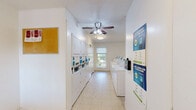 Located at 702 Edwards Rd in Greenville, South Carolina, this shared laundry facility offers a clean, functional, and well-organized space designed to serve residents efficiently. The room is laid out in a long, narrow configuration that maximizes the available floor area, with crisp white walls and durable tile flooring throughout that are easy to maintain and keep clean.

A row of commercial-grade top-load washing machines lines one side of the room, each equipped with modern digital payment interfaces for convenient, cashless transactions. Opposite the washers, stacked dryer units provide ample drying capacity. A ceiling fan with an integrated light fixture is centrally positioned overhead, promoting air circulation and comfort during use.

Natural light enters through windows fitted with blinds, creating a bright and welcoming environment. A second exterior door with a grid-pane window provides an additional access point and further enhances ventilation. Safety is addressed with a clearly mounted fire extinguisher near the entrance.

A cork bulletin board on the wall displays laundry tips and relevant notices for users. The space is rounded out by a waste receptacle for added convenience. Overall, this laundry facility is thoughtfully maintained and practically equipped to meet the day-to-day needs of its users.