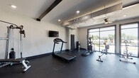 This fitness center offers a well-appointed, single-floor workout space designed with a clean, contemporary aesthetic. The interior features durable dark rubber flooring throughout, crisp white walls accented by bold black structural beams, and recessed lighting that keeps the space bright and functional. Exposed ductwork along the ceiling adds an industrial touch that complements the modern design language.

The cardio area is equipped with a treadmill, elliptical trainer, and a rowing machine, providing a comprehensive range of aerobic training options. A flat-screen television is wall-mounted for entertainment during workouts. The strength training section includes a multi-function cable machine, an adjustable weight bench, and a rack of dumbbells in varying weights, accommodating a wide range of resistance exercises.

A large floor-to-ceiling window and a sliding glass door flood the space with natural light and offer open views of the surrounding outdoor landscape, creating an airy, inviting atmosphere. A ceiling fan provides additional air circulation. The fitness center is accessed through glass-paneled double doors clearly labeled "Fitness Center," with a spin bike stationed just outside in an adjoining covered area. Interior windows connect the gym visually to an adjacent common area, enhancing the sense of openness and connectivity throughout the facility.