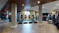 This expansive fitness center is designed to support a full range of workout routines within a thoughtfully organized, single-level layout. The space is anchored by a striking dark wood plank ceiling that lends warmth and sophistication, complemented by polished concrete floors and recessed lighting throughout.

The cardio zone features a row of treadmills equipped with individual screens, positioned in front of large floor-to-ceiling windows that invite natural light and provide outdoor views. Alongside the treadmills, a lineup of elliptical machines offers additional aerobic options. Ceiling fans ensure comfortable airflow across the open floor area.

The strength training section is well-appointed with a full dumbbell rack, adjustable benches, a power rack with barbell and weight plates, a cable multi-station, kettlebells, battle ropes, and a punching bag — catering to both functional and traditional weight training. Textured mosaic-tile accent walls define this zone, adding visual interest and an industrial edge.

A dedicated entry area features a built-in water station with a dual-level drinking fountain set against a natural wood and stone tile surround, accompanied by an upholstered bench with under-seat storage. Floor-to-ceiling mirrored walls in multiple areas enhance the sense of space and support form monitoring during workouts.