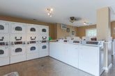 Located at 1040 Shive Ln in Bowling Green, Kentucky, this single-story community laundry facility is designed to meet the everyday needs of its users with an efficient and organized layout. The space features a generous number of commercial-grade top-loading washers and stacked dryers arranged along the walls, providing ample capacity for simultaneous use. The warm, neutral-toned walls with contrasting white wainscoting give the room a clean and welcoming ambiance. Durable tile flooring runs throughout, offering easy maintenance and a tidy appearance. Natural light filters in through windows fitted with vertical blinds, brightening the space and creating an open feel. Track lighting on the ceiling supplements the natural light, ensuring the room is well-lit at all times. A wall-mounted air conditioning unit maintains a comfortable temperature year-round. A central folding table provides a practical surface for sorting and folding laundry, while a bulletin board on the wall serves as a community information hub. A cushioned chair offers a comfortable spot to wait, adding a thoughtful touch to the functional space. The facility also includes a dedicated exit door for easy access and flow.
