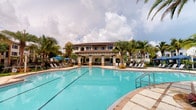 This inviting outdoor amenity area showcases a generous resort-style lap pool as its centerpiece, with clearly marked lanes and sparkling turquoise water that evokes a true vacation atmosphere. The pool is framed by a wide, tiled sun deck lined with lounge chairs, offering ample space for relaxation and sunbathing. Tall, mature palm trees are interspersed throughout the surrounding landscape, providing natural shade and enhancing the tropical ambiance of the setting. A covered outdoor pavilion anchors the poolside experience, supported by elegant columns and featuring what appears to be a social gathering and entertainment area. The pavilion's warm stucco facade and terracotta-tiled roof are characteristic of a Mediterranean or Spanish Colonial architectural style, adding a refined and cohesive aesthetic to the space. Blue market umbrellas provide additional shade near seating clusters, completing the resort-like feel. The backdrop features a multi-story building that shares the same architectural character, contributing to a unified and polished community design. The overall environment is bright, open, and well-maintained, making it an ideal outdoor destination for both leisure and social activity.