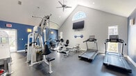 This fitness center is housed in a single-level space that makes a strong architectural impression with its dramatic vaulted ceilings, which rise to a striking peak and lend the room an open, airy feel. A distinctive arched window positioned high on the front wall floods the space with natural light, while recessed lighting and a ceiling-mounted fan ensure comfort during workouts. The interior is finished in a bold two-tone color scheme, with deep navy blue accent walls contrasting against crisp light gray surfaces, creating an energizing yet focused atmosphere. Durable dark rubber flooring runs throughout, providing a practical and professional surface underfoot. The fitness center is comprehensively equipped for a full-body workout experience. Cardio options include multiple treadmills, an elliptical trainer, a recumbent bike, and a WaterRower rowing machine. Strength training is well represented with a multi-station cable machine, a power rack loaded with Olympic weight plates, and additional resistance equipment. A wall-mounted flat-screen television adds an entertainment element for users during cardio sessions, and motivational wall decals reinforce the space's active ambiance. Mirrors along one wall enhance the sense of space and allow for proper form checks during exercise.